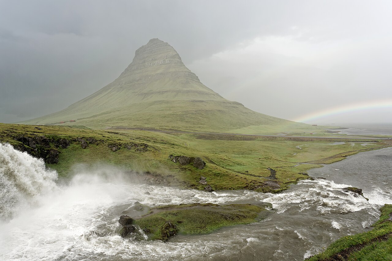 Kirkjufell rising behind Kirkjufellsfoss in rain and sunshine with a rainbow over the coast